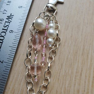 Puede incluir: Un llavero plateado con cierre de mosquetón, adornado con un grupo de cuentas y perlas de imitación. El diseño incluye cuentas facetadas rosas y transparentes, pequeñas cuentas rosas y perlas de imitación blancas de varios tamaños.