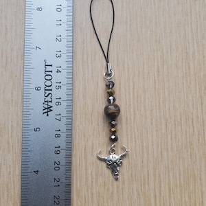 May include: A black and gold phone charm with a silver bull skull pendant and tiger's eye bead.