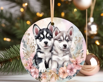 May include: A round Christmas ornament featuring a watercolor painting of two husky puppies surrounded by pink flowers. The ornament has a white background with a light pink and blue watercolor wash.
