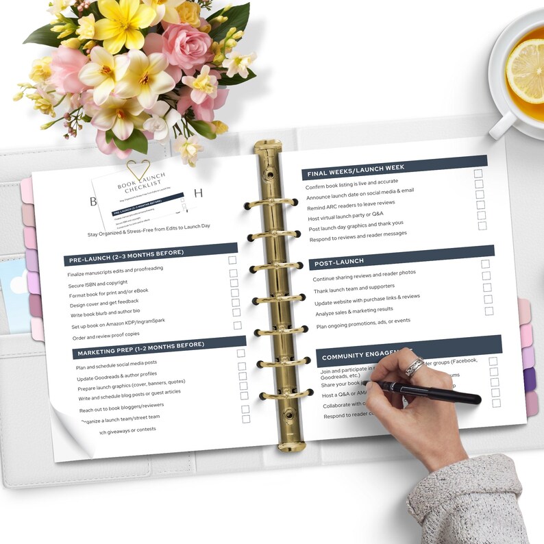 A flat lay of a planner with a book launch checklist. A hand holding a pen writes in the planner, surrounded by a bouquet of flowers and a cup of tea with lemon.
