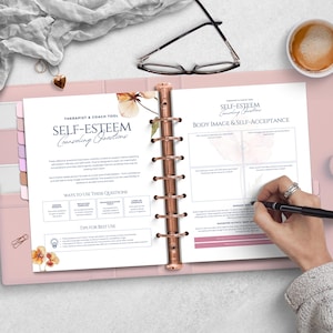 May include: An open pink planner with rose gold binding displays a page titled "SELF-ESTEEM Counseling Questions." The page includes sections on body image and self-acceptance. A hand with a pen is writing on the page. A coffee cup and glasses are on the table.