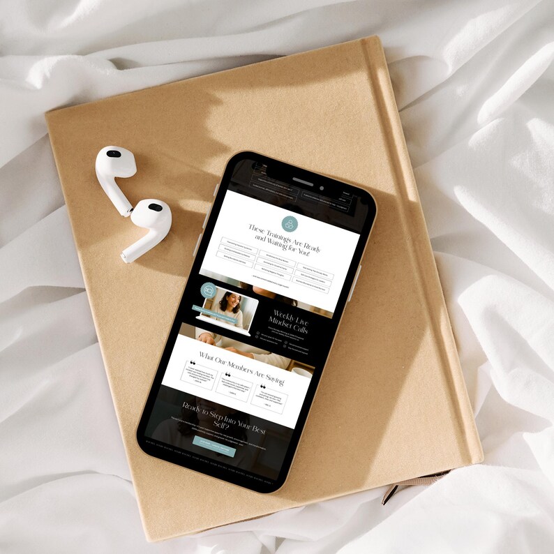 A smartphone displaying a webpage about training courses, placed on a tan notebook with white earphones beside it, resting on a soft white surface.