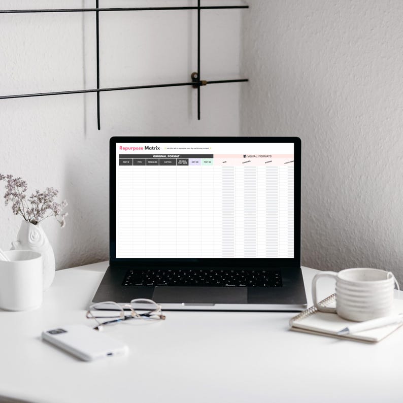 A laptop displaying a 'Repurpose Matrix' spreadsheet sits on a clean white desk with a coffee mug, glasses, and a notepad. Decorative elements include a small flower vase and a minimalist wall shelf.