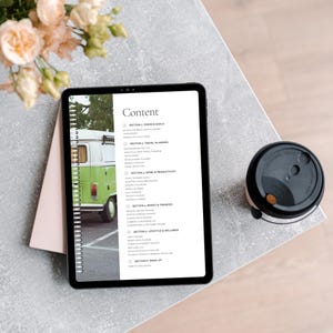 A tablet displaying a planner's content page, including sections like travel planning, work productivity, finance, and wellness. A pink notebook and a coffee cup are on a textured grey table, with flower arrangements in the background.