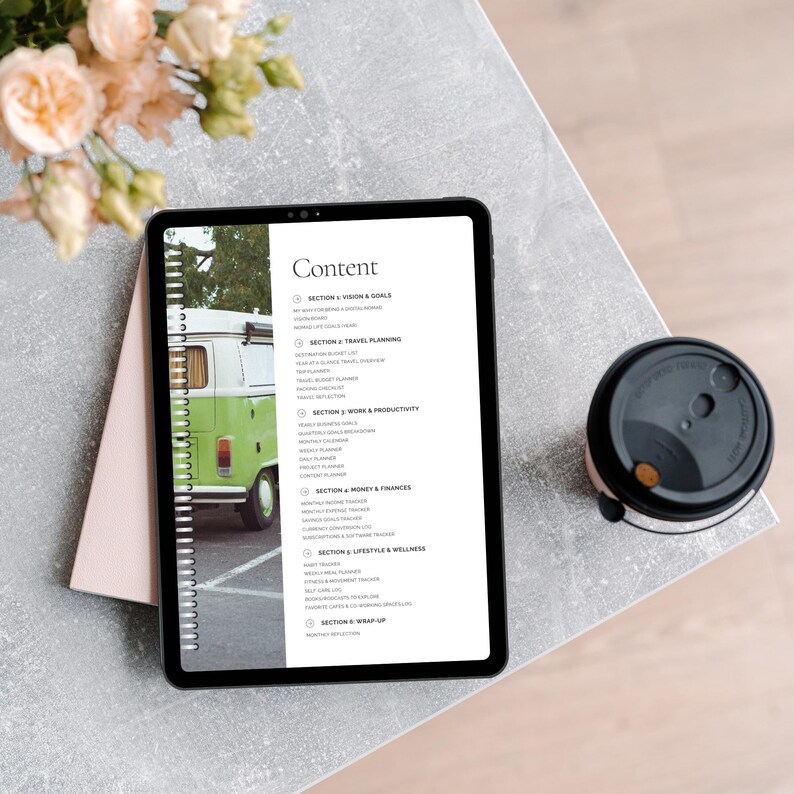 A tablet displaying a planner's content page, including sections like travel planning, work productivity, finance, and wellness. A pink notebook and a coffee cup are on a textured grey table, with flower arrangements in the background.