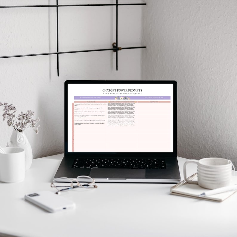 A desk scene featuring a laptop displaying a table titled 'ChatGPT Power Prompts for Marketing Your Business'. A white mug, a notebook, and glasses are on the desk. A small vase with flowers is also present.