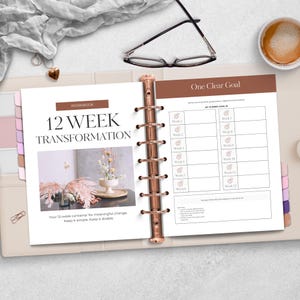 Op de afbeelding: Open planner met een 12-weekse transformatie werkboek. De omslag leest "12 WEEK TRANSFORMATION" met een bloemenafbeelding. De rechterpagina heeft een sectie voor een duidelijk doel en wekelijkse doeltracering. Een kopje koffie en een bril liggen op het witte oppervlak.