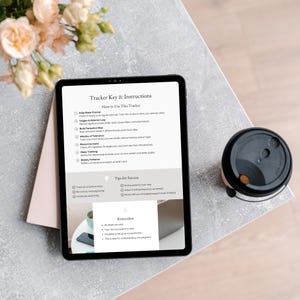 An iPad displaying a Tracker Key  Instructions sheet on a table, with tips for success and a reminder about mindfulness. A coffee cup is beside it, and there are soft blooms in the background.