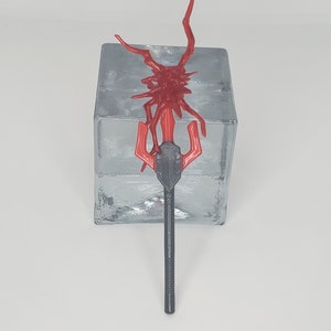 May include: A red and black spear-like object with a complex, spiky design at the top. The spear has a long, textured black handle and a red, trident-like head. The object is set against a textured, silver-colored background.