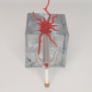 May include: A silver and red plastic toy sword with a red, spiky, starburst design on the top. The sword is on a clear glass block.