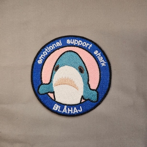May include: A blue and white embroidered patch with a cartoon shark and the text "emotional support shark" in a circle with a pink background. The text "BLÅHAJ" is below the shark.