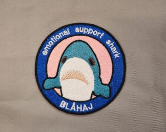 May include: An embroidered patch featuring a cartoon shark with the text "emotional support shark" in a circle with a pink background. The text "BLÅHAJ" is below the shark.