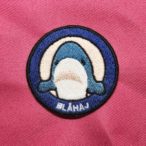 Puede incluir: Parche bordado azul y blanco de un tiburón de dibujos animados con el texto "BLAHAJ" en un círculo azul.