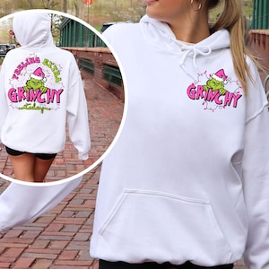 May include: White hoodie with a front pocket and drawstring hood. The front features a Grinch graphic with the word "Grinchy" in pink. The back of the hoodie has the text "Feeling Extra Grinchy Today" in a pink and green design.