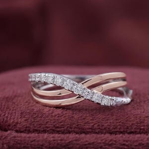 May include: A two-tone ring with a crisscross design. The ring features a band of small, sparkling white stones set in a silver-colored metal, crossing over a rose gold band. The ring is displayed on a burgundy velvet surface.