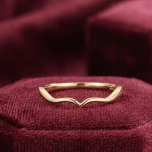 Puede incluir: Una alianza de boda dorada con un diseño curvo. El anillo tiene un acabado liso y pulido y una apariencia delicada. La banda se coloca sobre una superficie de terciopelo rojo oscuro.