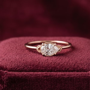 May include: A rose gold ring featuring a pear-shaped center stone held by a prong setting. The band includes small, detailed leaf-shaped accents on either side of the central stone, creating a delicate and refined appearance for this piece of jewelry.