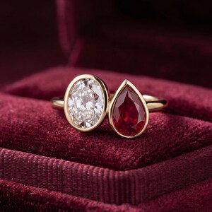 Pode incluir: Um anel de ouro com duas pedras em cravação inglesa: uma gema branca transparente de corte oval e uma gema vermelha profunda de corte em gota. O anel está posicionado sobre uma superfície de veludo vermelho escuro, exibindo uma banda polida.