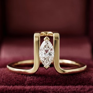 Può includere: Un anello d'oro con un diamante taglio marquise, incastonato tra due barre verticali d'oro. La fascia dell'anello si curva attorno al dito. L'anello è presentato su una superficie di velluto bordeaux, evidenziando il suo design elegante e la sua brillantezza.