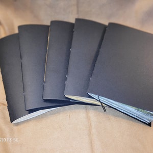 May include: Five black notebooks with sewn binding, stacked on a brown surface. The notebooks are closed and have a simple, minimalist design.