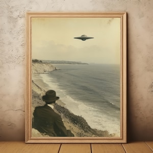 May include: Framed vintage-style print featuring a man in a suit and hat gazing at the ocean, with a UFO hovering in the sky. The sepia-toned image captures a coastal landscape with cliffs and waves.