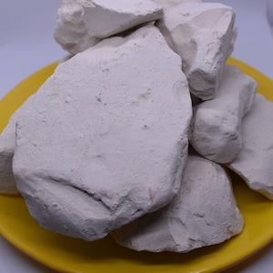 Puede incluir: Primer plano de un montón de rocas de arcilla blanca en un plato amarillo. La arcilla es de color gris claro y tiene una textura rugosa.