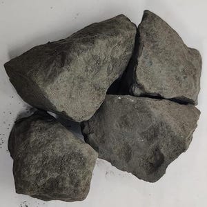 May include: Four dark grey rocks with irregular shapes are displayed on a white background. The rocks have a rough, textured appearance, with varying sizes and angular edges. The lighting creates subtle shadows, highlighting the uneven surfaces.
