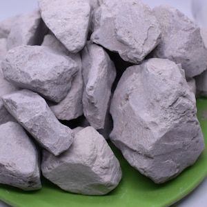 Può includere: Primo piano di un mucchio di rocce di argilla grigia chiaro su un piatto verde. Le rocce hanno una forma irregolare e una consistenza ruvida.