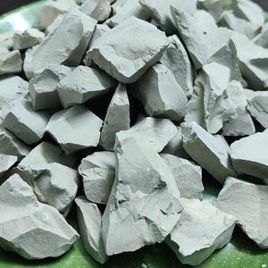 May include: A collection of light grey clay chunks, each with an irregular shape and a rough, fractured surface. The pieces vary in size and are piled together. The clay has a muted, neutral colour, suggesting a natural, raw material.