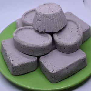May include: A stack of six lavender-coloured soap bars on a green plate. The bars are various shapes and sizes, including round, square, and rectangular.