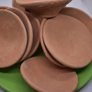 Puede incluir: Un conjunto de seis cuencos de terracota, todos con una forma y tamaño similares, están dispuestos sobre una superficie verde. Los cuencos están hechos de arcilla marrón rojiza y tienen un acabado liso y ligeramente texturizado.