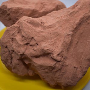 May include: Two large, irregularly shaped pieces of red clay on a yellow plate. The clay is soft and appears to be freshly dug.
