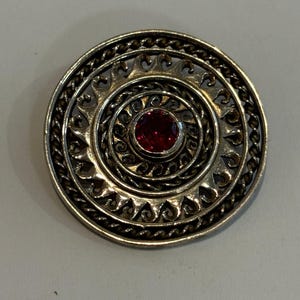 Könnte beinhalten: Runde, silberfarbene Brosche mit einem roten Edelstein in der Mitte. Die Brosche hat konzentrische Kreise mit aufwendigen filigranen Mustern und einem gedrehten, seilartigen Rand. Das Design ist symmetrisch und detailliert, zur Dekoration von Kleidung.