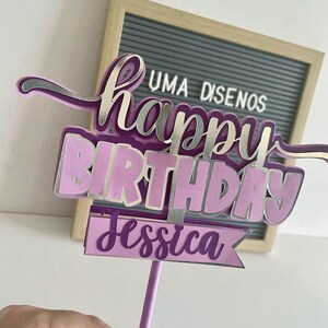 Hand holding a purple script Happy Birthday cake topper with layered details and a name tag reading Jessica on a purple stick.