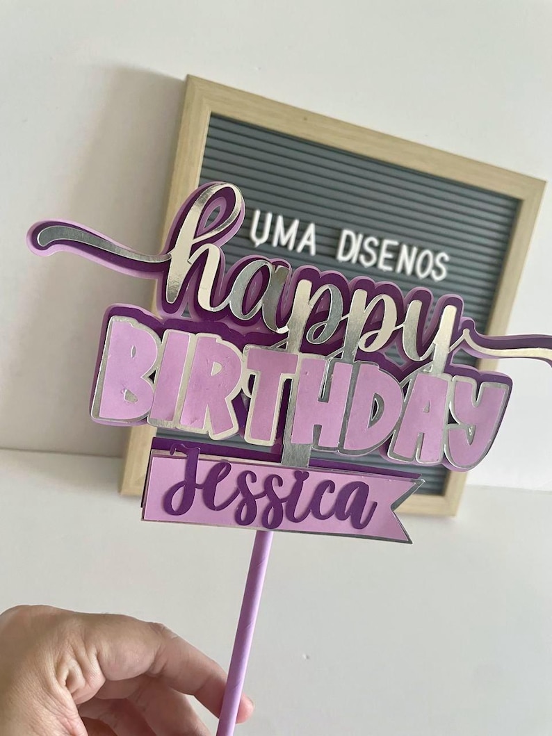 Hand holding a purple script Happy Birthday cake topper with layered details and a name tag reading Jessica on a purple stick.