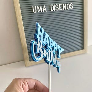 Side angle of a hand holding the blue 3D Happy Birthday cake topper, showing the multi-layered dimensional paper construction
