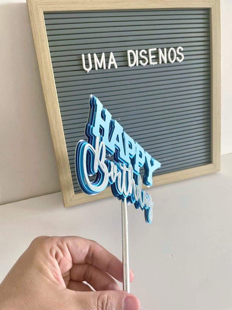 Side angle of a hand holding the blue 3D Happy Birthday cake topper, showing the multi-layered dimensional paper construction