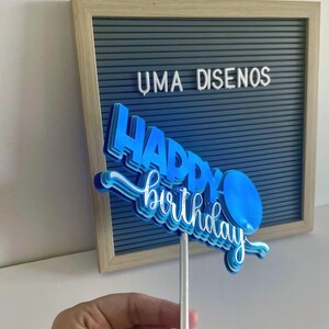 Hand holding a compact blue layered Happy Birthday cake topper on a stick centered in front of a gray felt letterboard.