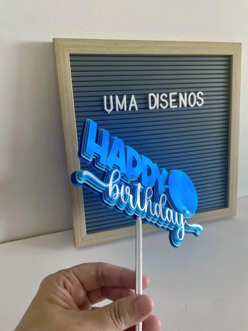 Hand holding a compact blue layered Happy Birthday cake topper on a stick centered in front of a gray felt letterboard.