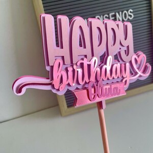 Hand holding a pink 3D Happy Birthday cake topper with a heart detail and a customizable name tag reading Olivia on a pink stick.