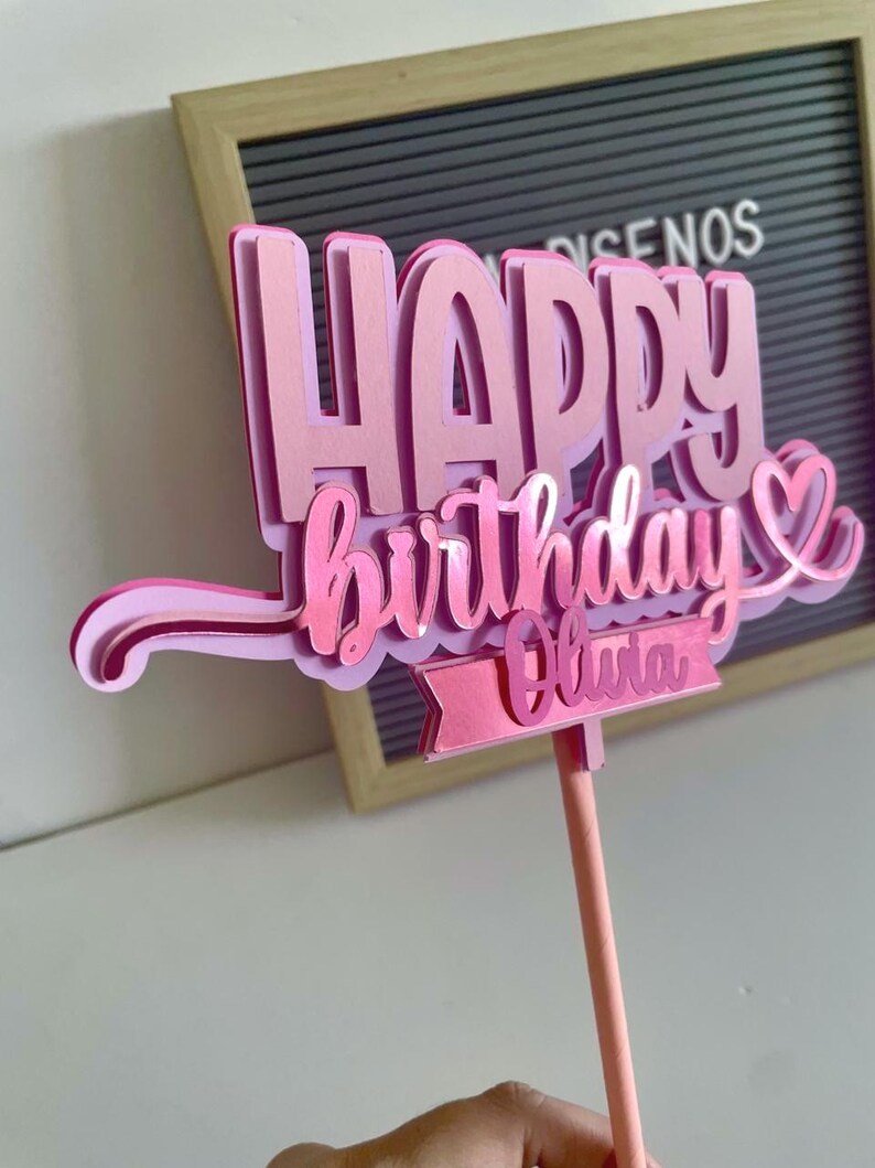 Hand holding a pink 3D Happy Birthday cake topper with a heart detail and a customizable name tag reading Olivia on a pink stick.