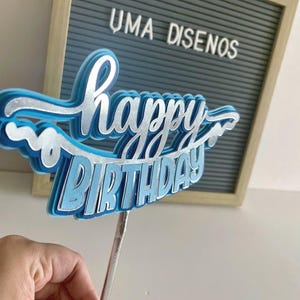 Hand holding a light blue banner-style Happy Birthday cake topper with white script text and wavy paper borders on a stick.