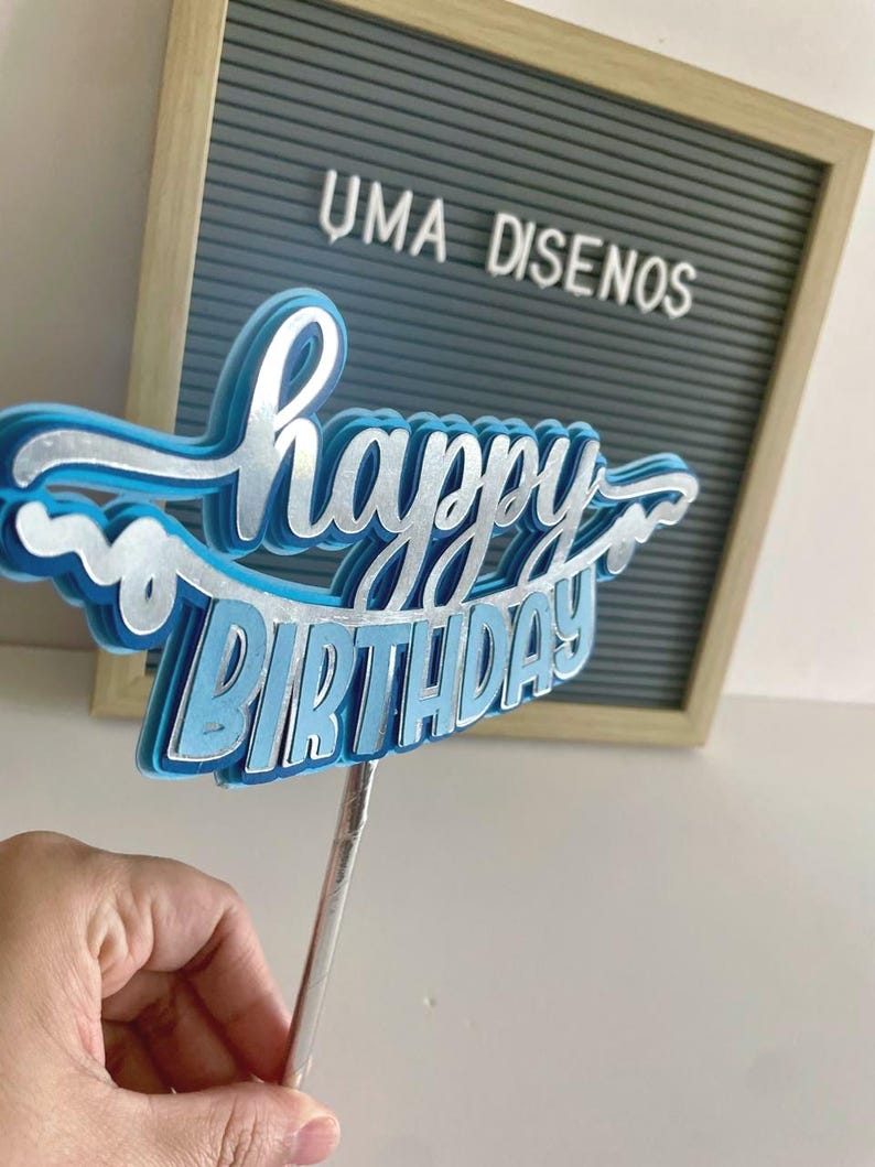 Hand holding a light blue banner-style Happy Birthday cake topper with white script text and wavy paper borders on a stick.