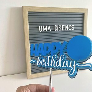 Hand holding a blue 3D Happy Birthday cake topper with an attached paper balloon accent on a stick.