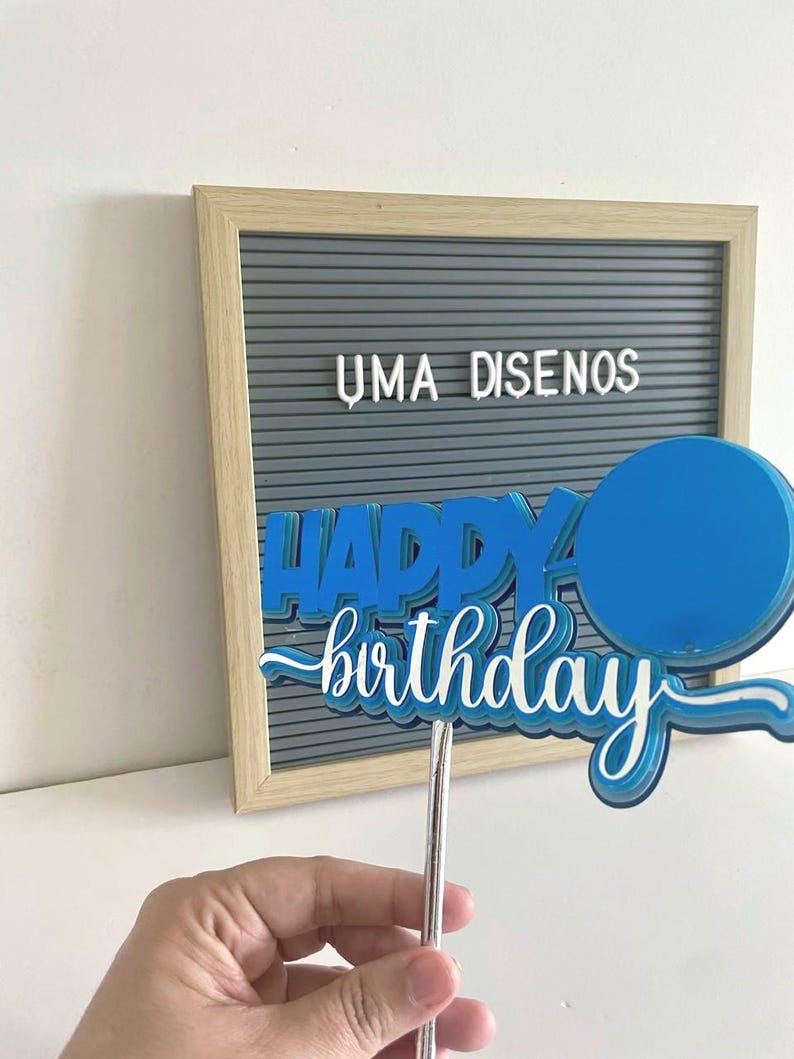 Hand holding a blue 3D Happy Birthday cake topper with an attached paper balloon accent on a stick.