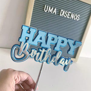 Hand holding a layered blue cardstock Happy Birthday cake topper on a stick in front of a gray wooden letterboard.