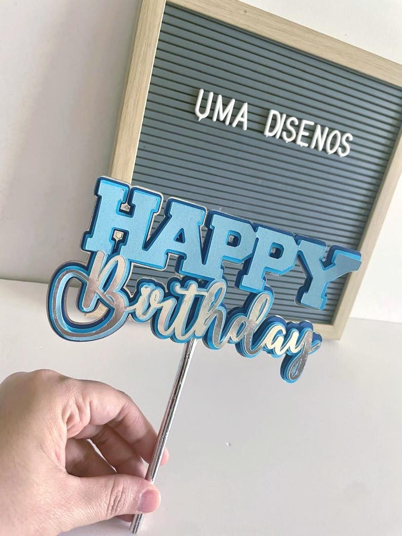 Hand holding a layered blue cardstock Happy Birthday cake topper on a stick in front of a gray wooden letterboard.