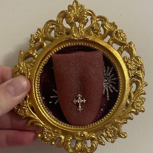 May include: A gold ornate frame with a red velvet interior, displaying a tongue with a small silver cross piercing. The tongue is surrounded by silver glitter.