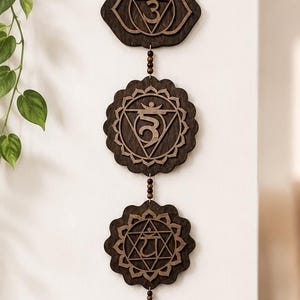 May include: A vertical wall hanging featuring seven dark brown wooden circular plaques, each laser-cut with a unique chakra symbol. The plaques are connected by small dark wooden beads and suspended from a single jute twine loop at the top.