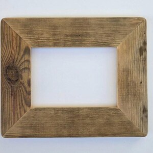 May include: A rectangular picture frame constructed from weathered brown wood with a visible grain pattern and a prominent knot on the left side. The frame features mitered corners and a wide, flat border surrounding a central rectangular opening.